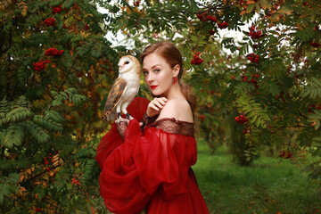 Beautiful red haired girl in red medieval dress with owl sitting on her hand among rowan. Closeup portrait of young woman with makeup - obrazy, fototapety, plakaty