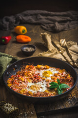 Shakshuka in an iron pan on wooden background. Middle eastern traditional dish. Fried eggs with tomatoes, bell pepper, vegetables and herbs. Vertical.