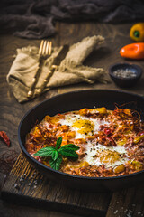 Shakshuka in an iron pan on wooden background. Middle eastern traditional dish. Fried eggs with tomatoes, bell pepper, vegetables and herbs. Vertical.
