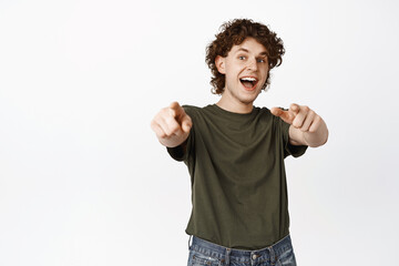 Its you. Excited young man pointing fingers at camera, shouting ehtusiastic, inviting or congratulating people, standing over white background