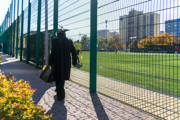 An old woman in a black coat with a black briefcase and autumn leaves in her hand walks past the fence of a football field along the sidewalk into the distance on a sunny autumn day