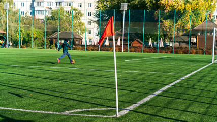 Red flag on the border of a green football field, long shadows of the fencing mesh in the rays of the evening sun, the figure of a child with a ball in the background