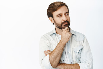 Thoughtful caucasian guy looking aside and touching his beard, thinking, making decision, standing over white background