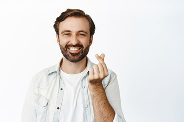 Cute bearded guy smiles and shows finger heart gesture, stands against white background