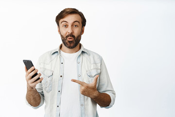 Amazed guy in casual clothes, pointing at telephone and looking impressed, showing interesting thing online, standing over white background
