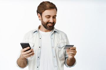 Handsome man enter credit card info in smartphone app, smiling pleased, standing over white background