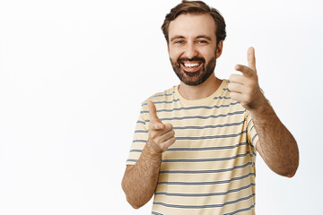 Image of joyful guy with beard, pointing fingers at camera and smiling, congratulating, praising and complimenting you, standing in t-shirt over white background