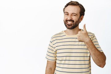 Handsome man winks and smiles, shows thumbs up in approval, recommends product, stands against isolated white background