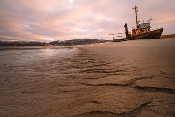 A rusty ship lies on a sandy shore at low tide