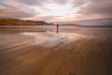 A man stands in the middle of the frame on a sandy beach at low tide. Seascape, red sky, reflection