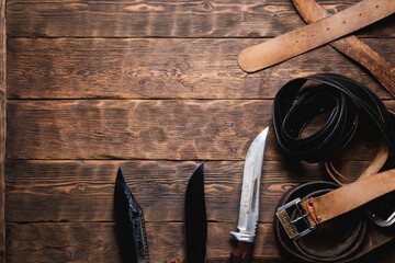 Hunting knives and old leather belts on the wooden flat lay board background with copy space.