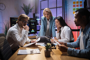 Female team leader in business clothes having briefing with multi ethnic colleagues at office room. Company workers sitting at desk, using modern gadgets and analysing financial statistics.