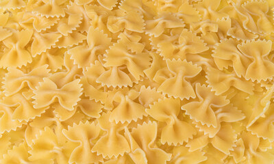 Uncooked dried bow-tie pasta as yellow culinary background. Close-up of raw egg farfalle from wheat flour dough. Beautiful texture from butterfly pastas heap. Foodstuff of Italian cuisine. Gastronomy.