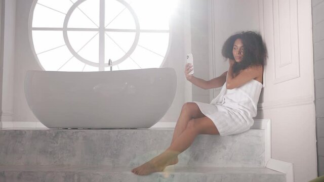 Black woman taking selfies on phone after bath, wrapped in towel Young afro girl after bath taking photos in bright white bathroom