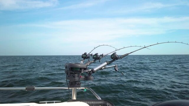Three fishing rods hooked up with spinners, rigged for fishing, with lines out in the water. The rods are lined up in a row on a small fishing boat. Gentle waves makes the boat rock. 