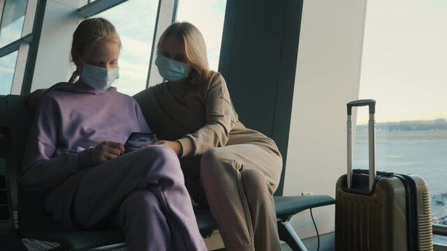 Mother and daughter waiting for their flight in the airport terminal. They are wearing protective masks, using a smartphone