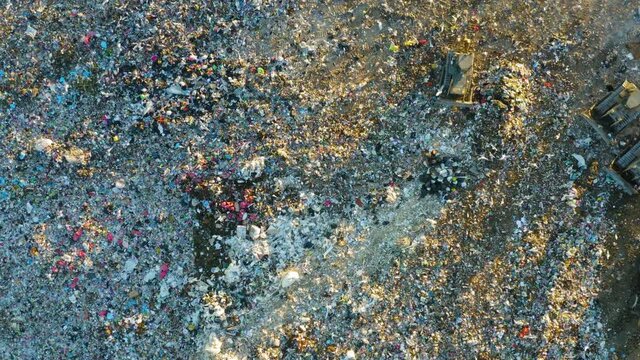 Aerial top down view of a city dump at sunset. Garbage trucks unload garbage. Bulldozers crush and level debris. Flocks of birds are circling over the dump.