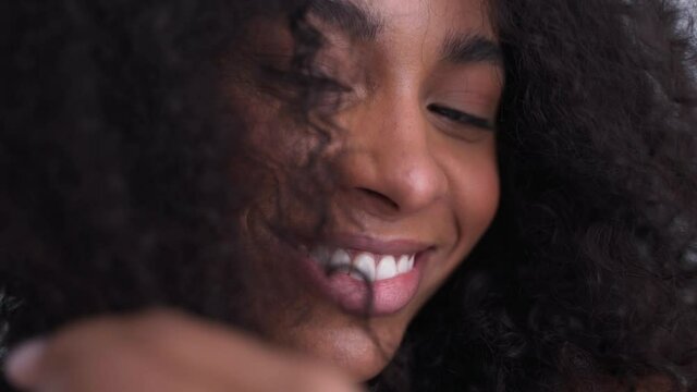 Close-up shot of black woman smiling at camera and playing with her afro hair in white room. Nature colours of woman hair routine. Dark skin girl smiling and playing with hair. High quality 4k footage