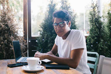 Portrait of intelligent male student in classic eyewear sitting in cafeteria with open terrace and...