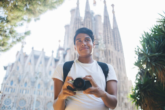 Carefree hipster guy with modern technology for clicking photos standing near Sagrada Familia and dreaming about getaway, happy male tourist holding vintage camera enjoying international trip