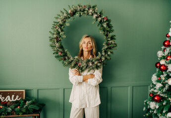 Conceptual portrait of young elegant woman framed by christmas wreath against wall in living room - Powered by Adobe