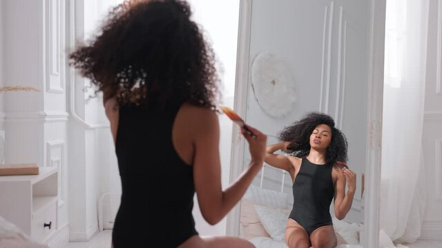 Black woman combing her afro hair, sitting on a bed and looking in mirror. Taking hair care of afro hair in bright white bedroom. High quality 4k footage