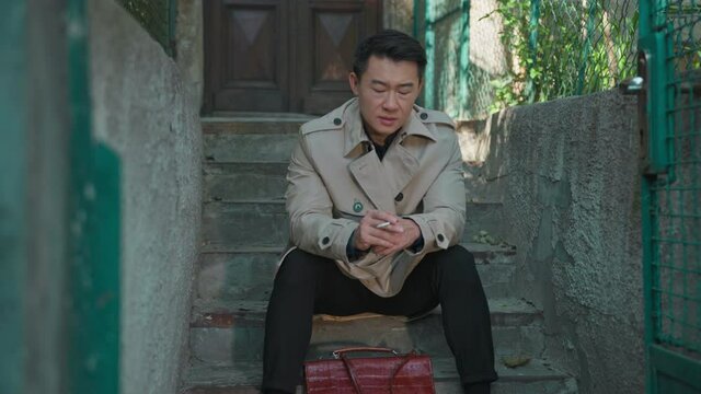Chinese frustrated businessman thinking about loss sitting on stairs relaxing outside and smoking cigarette. Failure. Frustration. Emotional people. Stress.