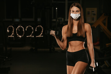 A fit woman in a face mask to avoid the spread of coronavirus is doing bicep curls with dumbbells....