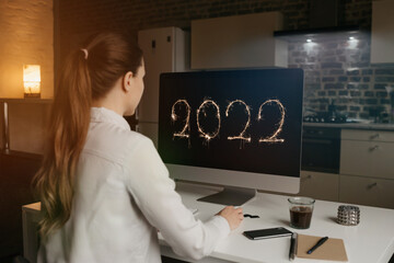 A back view of a woman who is staring at the number 2022 on a display of her desktop computer at home. A businesswoman is watching a new year video.