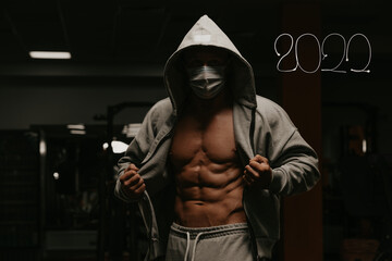 A bodybuilder in the hood and a mask to avoid the spread of coronavirus is opening his zipped hoodie to demonstrate his abs. A sporty guy in a surgical mask is posing after a workout in a gym. 2022