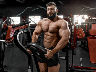 Brutal man with a beard holds a 25 kg disc in his hands in the gym. A muscular bodybuilder in perfect athletic shape is preparing for a workout on a sports simulator