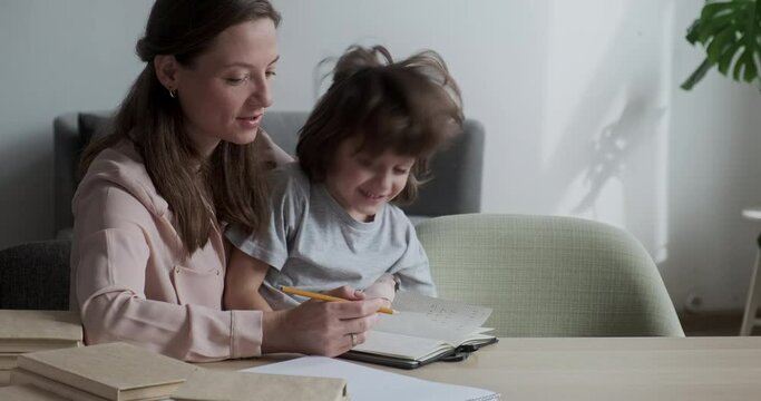 Cute smiling boy fooling playing at desk and having fun, mother having hard time with learning and homework. Hyperactivity in preschoolers and parental support and care.