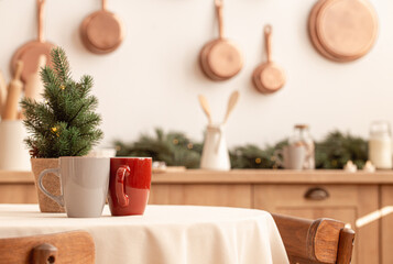 Two tea cups on Christmas table with little tree in bright modern decorated kitchen interior with many copper pans on wall, kitchen tools and utensils, candles, space for text
