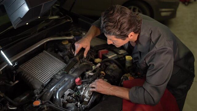 High angle view of the auto mechanic standing at the opened hood and lighting with lamp while working at the workshop and repairs injector. Automotive electrician concept