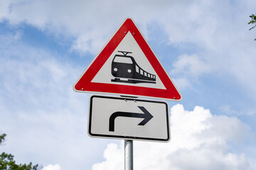 Rail crossing without barriers ahead traffic sign in Germany. Warning symbol to pay attention in front of the sky. A black train on a white triangle with a red outline. An arrow pointing to the right.