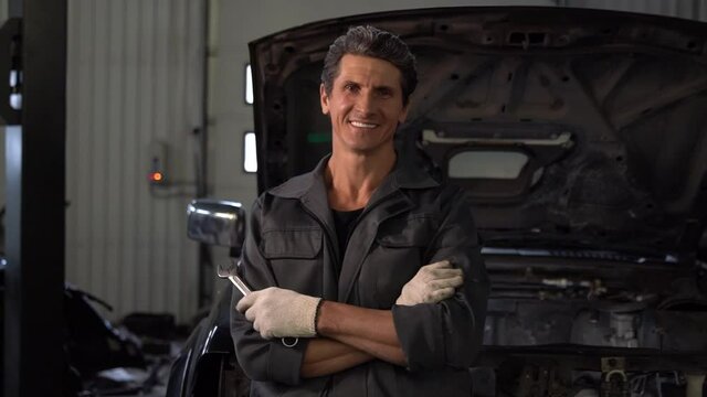 Waist up portrait view of the senior auto mechanic posing arms crossed with wrench at workshop and smiling toothy to the camera. Car service and repair concept