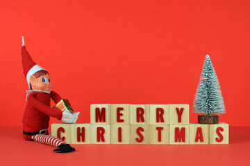 Merry Christmas written on wooden blocks with a Christmas tree and festive elf on a red background.