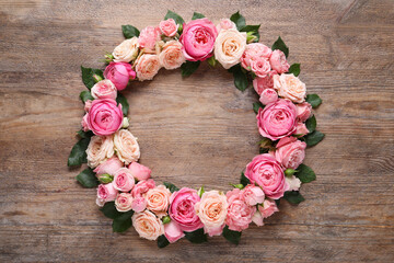 Wreath made of beautiful rose flowers and green leaves on wooden background, flat lay. Space for text