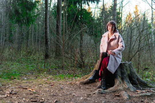 A girl sits on a stump in a coniferous forest in a red dress over a pink polto