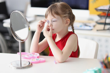 Little girl painting her eyes with baby makeup in front of mirror