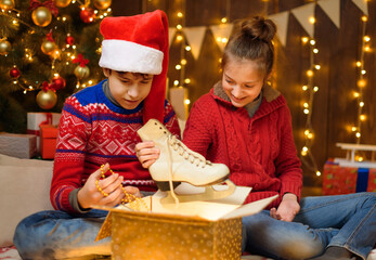 Fototapeta na wymiar Portrait of a boy and girl in New Year decoration. They open a box, take out their skates and have fun. Holiday lights, gifts and a Christmas tree decorated with toys.