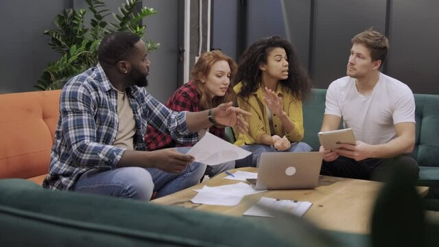 Positive multicultural young people in casual wear laughing while discussing collaborative plan sitting at table with laptop computer. Teamwork of diverse guys during brainstorming
