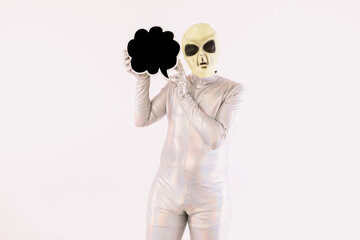 Person dressed in silver suit and green alien mask, holding a comic speech bubble with hand, on...
