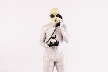 Person dressed in silver suit and green alien mask, talking on a black retro telephone from the 60s, on white background
