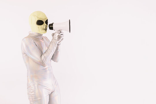 Person dressed in silver suit and green alien mask, speaking with a white megaphone, on white background