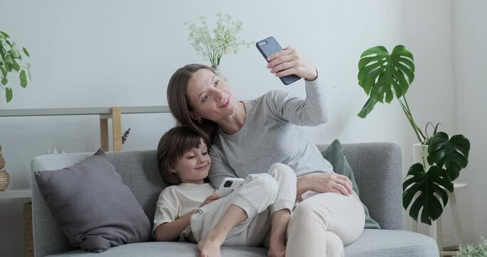 Smiling mother and cute little son posing taking selfie use smartphone relaxing couch enjoying family weekend. Happy woman and kid boy photographing together having positive emotion at home interior