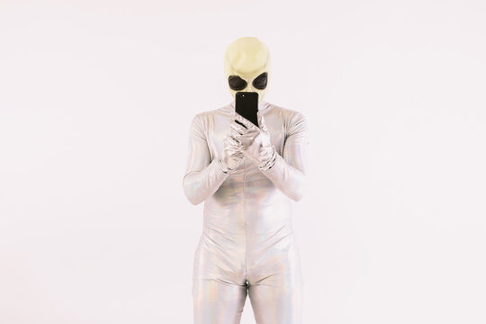 Person dressed in silver suit and green alien mask looking at mobile phone over white background