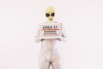 Person dressed in silver suit and green alien mask, holding an Area 51 poster, on white background