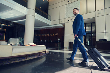 Handsome man, successful business developer smiling, looking to the side, walking down the lobby, heading with luggage to the lounge area inside a modern luxury building during at trip © Taras Grebinets