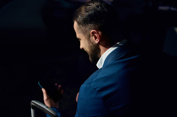 Rear view of a well-groomed attractive handsome bearded businessman in business casual suit using a mobile phone while sitting on a chair in a hotel lounge during his business trip. Copy space